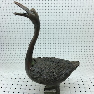 Puede incluir: Una figura de pato de bronce con pico abierto y textura detallada de plumas. El pato tiene un cuello largo y curvado y patas palmeadas. Es un objeto decorativo.