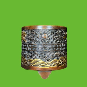 Peut inclure: Un récipient cylindrique en métal de couleur bronze orné de reliefs complexes représentant des nuages tourbillonnants, des motifs géométriques et des vagues stylisées. La surface présente une patine sombre avec des accents dorés.
