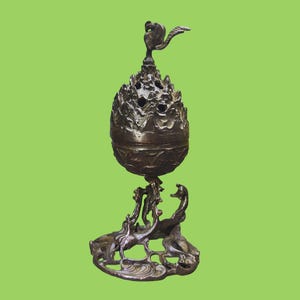 May include: A dark bronze-colored metal incense burner featuring a spherical, perforated top section with a small bird-shaped finial. The base is composed of intricate, openwork metal designs depicting swirling patterns and a small bird figure.