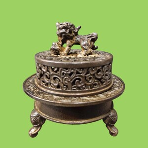 May include: A dark metallic incense burner featuring a circular base with three legs and a removable lid. The lid is decorated with intricate openwork patterns and topped with a sculpted figure of a lion-like creature resting on its paws.