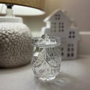 May include: A clear, cut-glass jar with a lid, featuring a diamond pattern. The jar is small and sits on a white surface. A decorative white house and a lamp are in the background.
