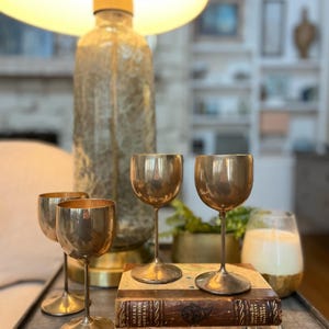 Puede incluir: Una colección de copas de vino antiguas de latón con tallos delgados, dispuestas sobre una superficie oscura con libros antiguos. Una lámpara grande y ornamentada y una vela en un tarro de vidrio están en el fondo.