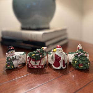 Op de afbeelding: Een verzameling van vier keramische servetringen met kerstthema's. De ontwerpen omvatten een sneeuwpop, een kerstboom op een slee, de kerstman en een kerstboom. Elke ring is wit met kleurrijke accenten.