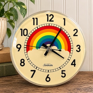 May include: A round Sunbeam analog clock with a cream-colored face, black numerals, and black hands. The center features a colorful rainbow graphic arching over a white cloud. The clock has a clear circular frame and a simple, clean design.
