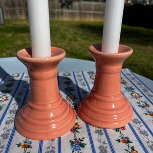 May include: Two coral-colored ceramic candle holders with white candles. The candle holders have a fluted design and are set on a floral patterned tablecloth. The background is a sunny outdoor scene.