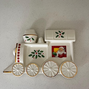Puede incluir: Una fuente de cerámica blanca con forma de tren navideño con ribete dorado. El plato presenta una ilustración de Papá Noel, detalles de acebo y detalles rojos y dorados. Las ruedas del tren son blancas con bordes dorados.