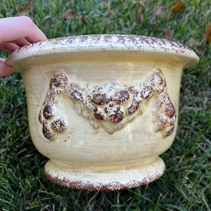 May include: A cream-colored ceramic planter with a decorative brown floral design. The planter has a wide, rounded shape with a pedestal base. The rim and base have a distressed, textured appearance, adding to its vintage charm. Perfect for indoor or outdoor use.