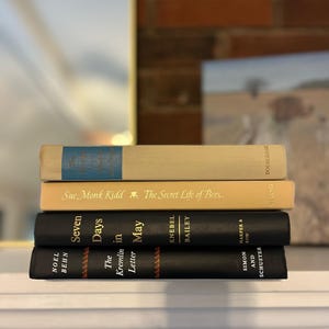 May include: A stack of four hardcover books with varying colors and titles. The top book is light beige with a blue label. The second book is beige with gold lettering. The bottom two books are black.