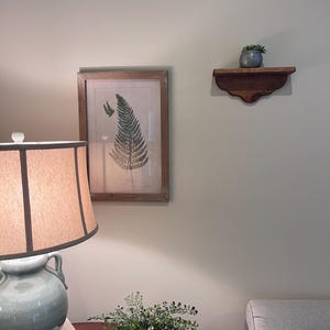 May include: A framed botanical print of a fern hangs on a light-colored wall. A wooden shelf holds a small potted plant. A table lamp with a beige shade and a ceramic base sits on a table with a decorative plant.