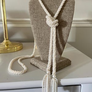May include: A long lariat necklace crafted from small white seed pearls woven into a rope-like texture. The necklace features a central decorative knot and two dangling ends finished with matching pearl tassels, creating a continuous strand design.