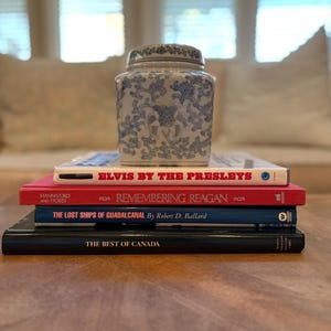 Puede incluir: Una pila de libros con un tarro de cerámica con estampado azul y blanco encima. Los libros incluyen títulos como "Elvis by the Presleys", "Remembering Reagan", "The Lost Ships of Guadalcanal" y "The Best of Canada".