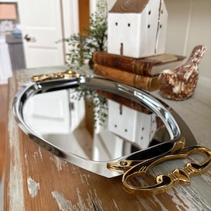 May include: A polished, oval-shaped silver serving tray with gold-colored handles. The tray reflects the surrounding objects. Decorative gold-colored scissors are placed on the wooden surface. A small white house figurine and a bird figurine are also visible.
