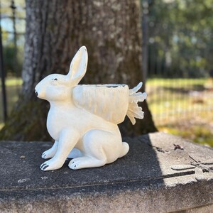 Puede incluir: Una maceta blanca con forma de conejo y acabado envejecido. El conejo está sentado con las orejas erguidas. El área de la maceta está ubicada en la espalda del conejo, diseñada para contener pequeñas plantas o flores. La maceta está sobre un banco de piedra.