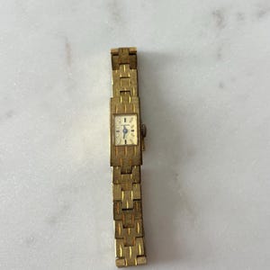 May include: A vintage gold-coloured wristwatch with a rectangular face and a linked band. The watch face is a light colour with black and blue hands. The band has a textured, geometric pattern. The watch is approximately 16.5 cm long.
