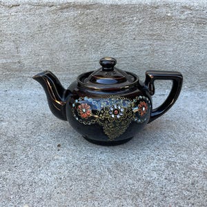 Puede incluir: Una tetera de cerámica marrón oscuro con tapa y asa. La tetera está decorada con un diseño floral en naranja, blanco y dorado. La tetera está sobre una superficie de hormigón.