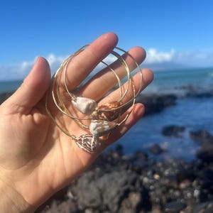 May include: Four gold-colored bangles, each adorned with a unique seashell. The shells display a mix of white, brown, and tan hues. The bangles are held against a backdrop of a blue sky and ocean.
