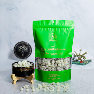 Puede incluir: Una bolsa verde brillante de incienso Omani Hojari, con una ventana transparente que muestra el producto. La bolsa está etiquetada con texto en inglés y árabe. Un pequeño cuenco negro y dorado con incienso se encuentra sobre un soporte de madera.