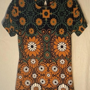 Op de afbeelding: Kort mouw jurk met een levendig bloemenpatroon in oranje, wit, teal en zwart. De jurk heeft een sleutelgatrug en een getailleerd silhouet. Het ontwerp bevat grote gestileerde bloemen en wervelende details.