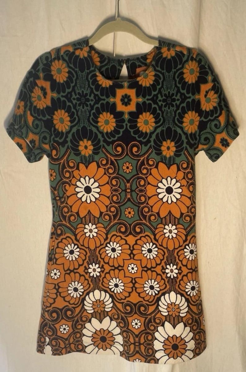 Op de afbeelding: Kort mouw jurk met een levendig bloemenpatroon in oranje, wit, teal en zwart. De jurk heeft een sleutelgatrug en een getailleerd silhouet. Het ontwerp bevat grote gestileerde bloemen en wervelende details.