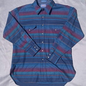 May include: A long-sleeved button-down shirt with a striped pattern in shades of blue, teal, and burgundy. The shirt has two chest pockets, a collar, and buttoned cuffs. The brand name is visible on the collar.
