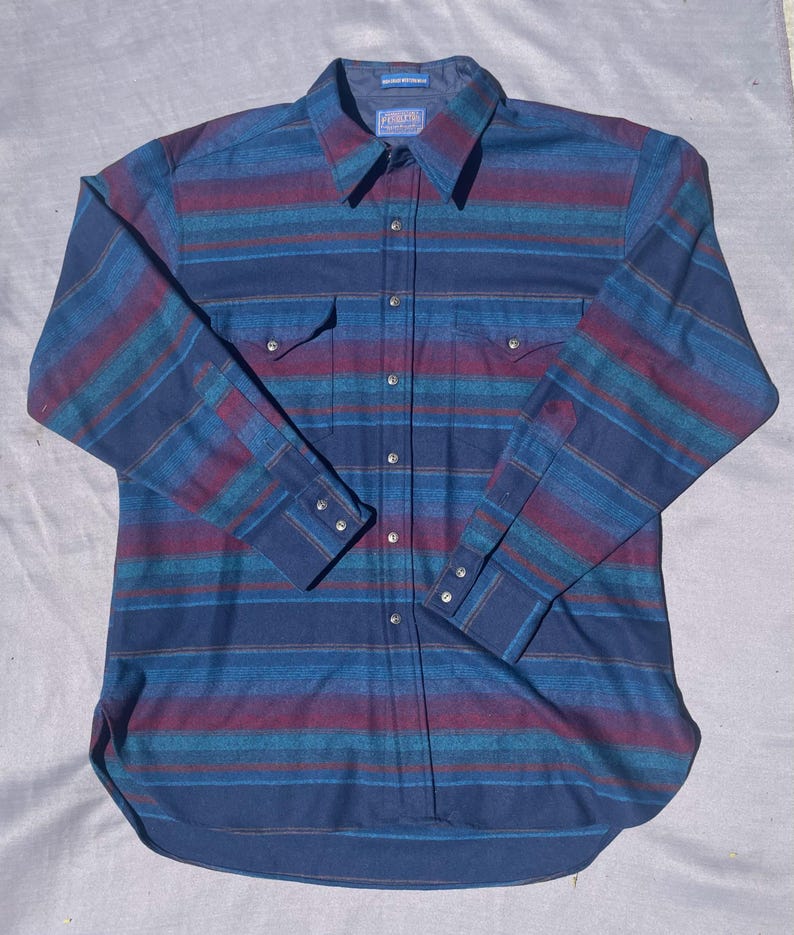 May include: A long-sleeved button-down shirt with a striped pattern in shades of blue, teal, and burgundy. The shirt has two chest pockets, a collar, and buttoned cuffs. The brand name is visible on the collar.