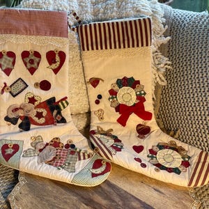 Puede incluir: Dos calcetines navideños de tela color hueso con texturas acolchadas, ribetes de encaje y aplicaciones de tela variadas que incluyen corazones, estrellas y figuras. El calcetín izquierdo tiene un puño de cuadros rojos y el derecho rayas.