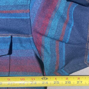 May include: Close-up of a blue, teal, and burgundy striped shirt. The shirt has a button-down front and a pocket. The fabric appears to be a woven material. The shirt is measured with a tape measure.