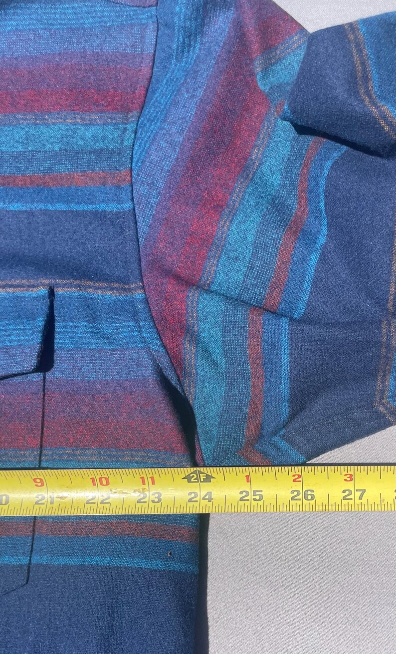 May include: Close-up of a blue, teal, and burgundy striped shirt. The shirt has a button-down front and a pocket. The fabric appears to be a woven material. The shirt is measured with a tape measure.