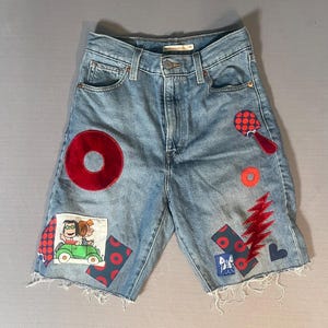 May include: Light blue denim shorts with a high waist and frayed hems. The shorts feature various fabric patches including a red velvet circle, a red lightning bolt, a cartoon illustration of two characters in a green car, and red polka-dot patterns.
