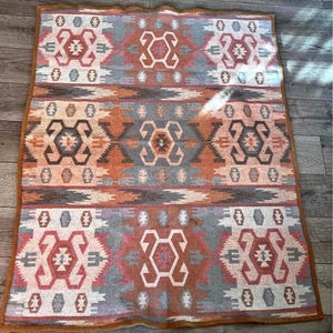 May include: A rectangular woven blanket with a Southwestern geometric pattern. The blanket features a repeating design in shades of red, orange, gray, and white. The blanket has a brown border and is likely made of a soft material.