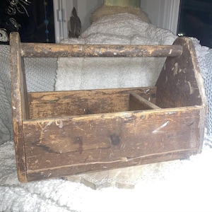 May include: A rustic wooden toolbox with a handle, showing signs of age and wear. The box is rectangular with a divided interior, suitable for storing tools or small objects. The wood has a distressed finish, with visible grain and imperfections.