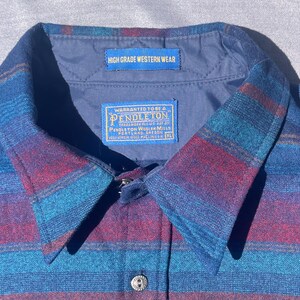 May include: Close-up of a blue, red, and teal striped button-down shirt. The collar features a label that reads "HIGH GRADE WESTERN WEAR." Another label reads "WARRANTED TO BE A PENDLETON." The shirt has a button closure and a single button on the collar.