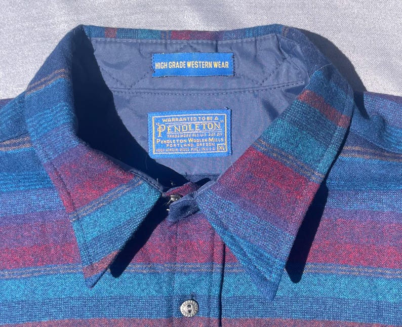 May include: Close-up of a blue, red, and teal striped button-down shirt. The collar features a label that reads "HIGH GRADE WESTERN WEAR." Another label reads "WARRANTED TO BE A PENDLETON." The shirt has a button closure and a single button on the collar.