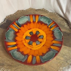May include: A round, decorative plate with a vibrant floral design. The plate features a deep red border, with a central pattern of orange, yellow, and teal shapes. The plate is placed on a wooden tray with a decorative edge.
