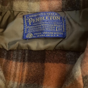 May include: Detail of a Pendleton wool jacket label. The blue label states "WARRANTED TO BE A PENDLETON" with the Pendleton Woolen Mills logo and "100% VIRGIN WOOL." The jacket features a brown, orange, and tan plaid design.