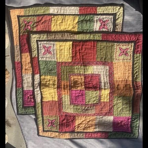 May include: Two colorful quilted placemats with a patchwork design. The squares are in shades of red, yellow, green, and pink. Each placemat has a floral embroidered accent and a dark brown border.