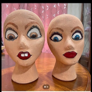 May include: Two ceramic mannequin heads with painted faces. One head has brown eyes, a red mouth, and a grumpy expression. The other head has blue eyes and a red mouth with a neutral expression. Both heads are on a wooden table.