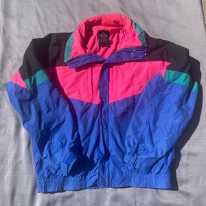 May include: A vintage Columbia jacket in a vibrant color block design. The jacket features a combination of bright pink, blue, black, and teal. It has a high collar, a zipper closure, and elastic cuffs. The Columbia logo is visible on the collar.
