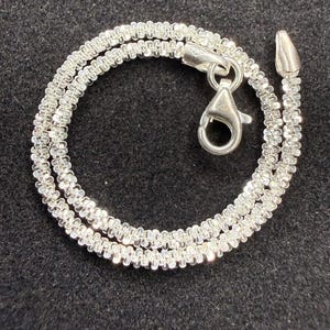 Könnte beinhalten: Eine silberfarbene Popcorn-Kette mit einem strukturierten Design aus mehreren Gliedern. Die Kette wird mit einem polierten Karabinerverschluss und einer passenden Endkappe gesichert, was eine durchgehende, flexible Schlaufe aus Metallgliedern bildet.