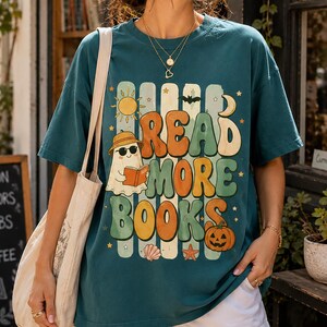 May include: A dark teal short-sleeve t-shirt featuring a graphic print with the text READ MORE BOOKS. The design includes a ghost wearing a hat and sunglasses, a pumpkin, a sun, a moon, a bat, a seashell, and a starfish in muted colors.