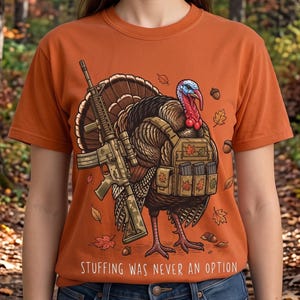 May include: An orange t-shirt featuring a graphic of a turkey wearing a tactical vest and holding a rifle. The turkey is surrounded by autumn leaves and acorns. The text "STUFFING WAS NEVER AN OPTION" is printed in white at the bottom of the design.