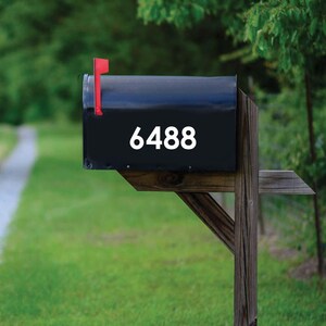 May include: A black mailbox with the number 6488 in white. The mailbox has a red flag and is mounted on a wooden post. The background is a blurred view of green grass and trees.