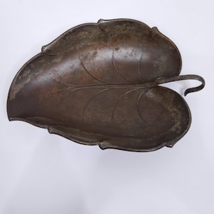May include: A leaf-shaped metal tray with a handle. The tray has a dark, aged appearance with visible veining details, and a slightly curved handle. The overall design suggests a decorative or serving piece, possibly for a rustic or vintage aesthetic.