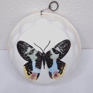 Könnte beinhalten: Weiße Keramik-Wanddekoration mit Schmetterlingsdesign. Der Schmetterling hat schwarze, blaue und gelbe Flügel. Ein Metallring ist zum Aufhängen angebracht. Das Stück hat einen gewellten Rand und dient wahrscheinlich dekorativen Zwecken.