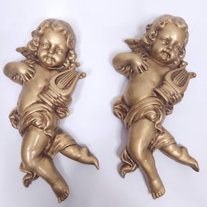 May include: Two gold-coloured cherub wall sculptures. Each cherub is depicted with wings, holding a harp, and draped with fabric. The sculptures have a classical, antique aesthetic and are set against a white background.