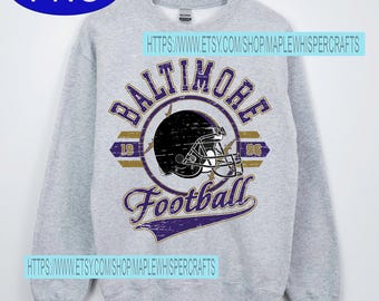 Può includere: Una felpa grigio chiaro con una grafica di casco da football nero e la scritta "Baltimore Football" in viola e oro. La felpa ha maniche lunghe e girocollo. Viene visualizzato anche l'anno 1996.