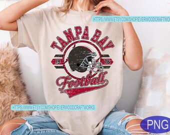 Puede incluir: Camiseta beige con un gráfico desgastado. Presenta un casco de fútbol dentro de un círculo, con las palabras "TAMPA BAY Football" en rojo. La camiseta también incluye los números "19" y "76".