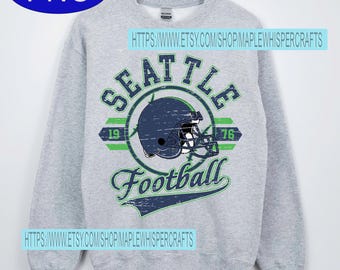 May include: A gray sweatshirt featuring a distressed design with the text "SEATTLE Football" and a football helmet graphic. The design includes the years "19" and "76" in green. The sweatshirt is displayed on a wooden hanger.