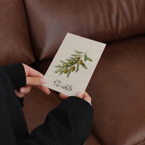 May include: A rectangular off-white card featuring a watercolor illustration of an olive branch with green leaves and olives. Below the illustration, the card displays the Arabic text "والزيتون" enclosed in decorative brackets with the number one.