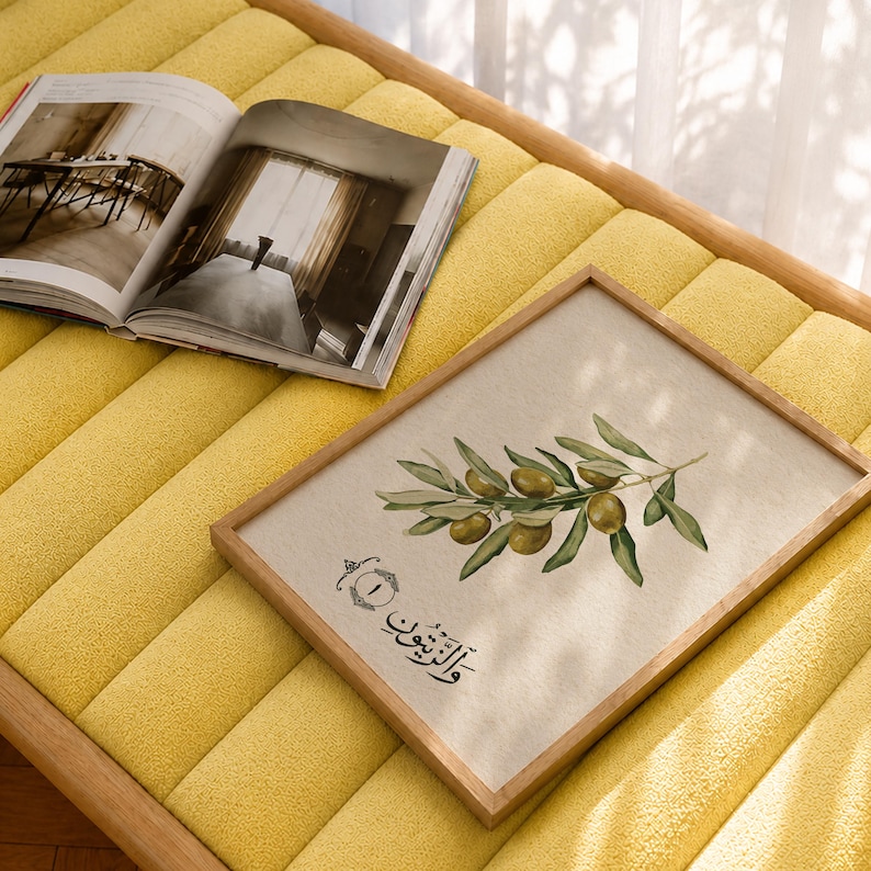 May include: A rectangular wooden frame containing a print of an olive branch with green olives and leaves, featuring Arabic calligraphy and the number one in a decorative circle. An open book with black and white interior photography is nearby.